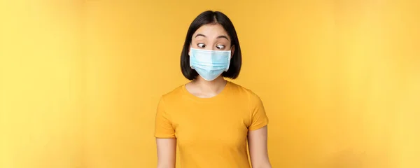Conceito de saúde e pessoas. Engraçado asiático menina squinting, olhando para seu rosto máscara médica, de pé sobre fundo amarelo — Fotografia de Stock