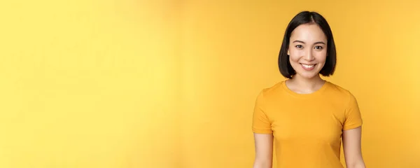 Portret młodej nowoczesnej Azjatki, uśmiechnięta szczęśliwa z białymi zębami, pewnie patrząca w kamerę, ubrana w luźny t-shirt, stojąca nad żółtym tłem — Zdjęcie stockowe