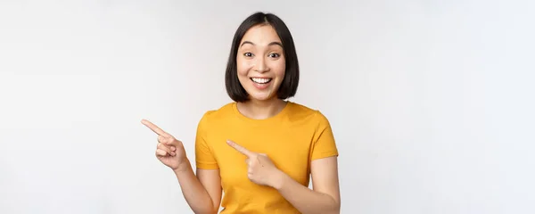 Портрет улыбающейся азиатской брюнетки в желтой футболке, указывая пальцами влево, показывая пространство для копирования, рекламную сделку, демонстрируя баннер, стоя на белом фоне — стоковое фото