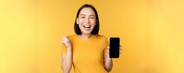 Excited asian woman, showing smartphone app and triumphing, celebrating on mobile phone, standing over yellow background — Stock Photo, Image