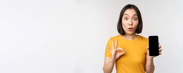 Spännande asiatisk flicka som visar mobiltelefon skärm, okej skylt, rekommendera smartphone app, står i gul t-shirt över vit bakgrund — Stockfoto