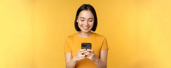 Технології. Усміхнена азіатська жінка, використовуючи мобільний телефон, тримає смартфон в руках, стоячи в футболці на жовтому тлі — стокове фото