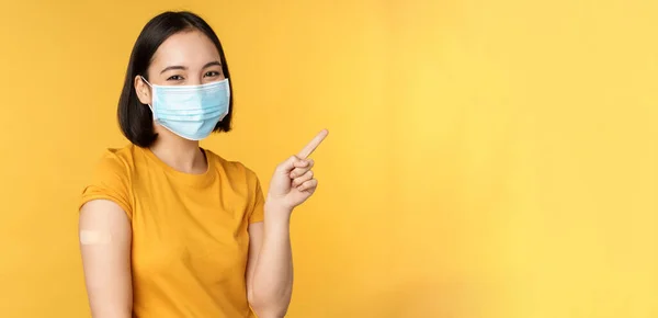 Vaccination från covid och hälsa koncept. Bild av leende koreanska flicka i medicinsk ansiktsmask, band stöd på axeln, peka finger på banner annons, gul bakgrund — Stockfoto