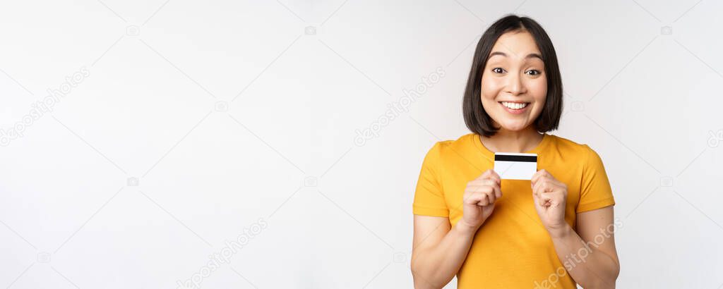 Portrait of beautiful korean girl holding credit card, recommending bank service, standing in yellow tshirt over white background