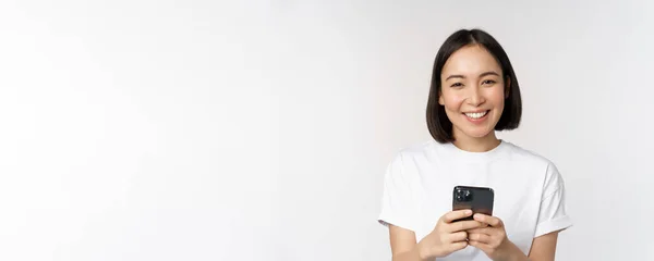 Retrato de sorrindo mulher asiática usando telefone celular, bate-papo, mensagem de texto, de pé em tshirt sobre fundo branco — Fotografia de Stock