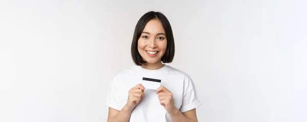 Sorridente coreano donna mostrando carta di credito con volto felice, in piedi in t-shirt su sfondo bianco — Foto Stock