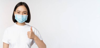 Tıbbi maskeli gülümseyen Asyalı kız başparmağını kaldırıyor, iyi bir şey onaylıyor, övgü ve iltifat şirketi beyaz arka planda duruyor.