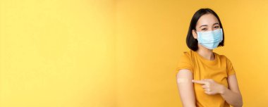 Aşı ve covid-19 pandemik konsept. Yüzünde tıbbi maske olan gülümseyen Asyalı kadın. Koronavirüs aşısı sonrası omzunu yara bandıyla gösteriyor. Sarı arka plan.