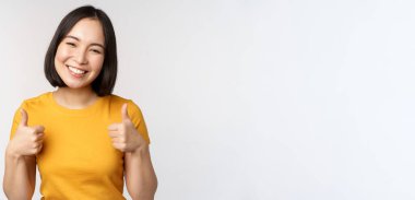 Güzel gülümseyen Asyalı kadın model, başparmaklarını kaldırıyor ve memnun görünüyor, tavsiye ediyor, olumlu geri bildirim yapıyor, beyaz arka planda duruyor.
