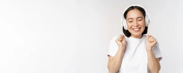 Moderne asiatique fille danse, écouter de la musique avec des écouteurs, souriant heureux, debout en t-shirt sur fond blanc — Photo