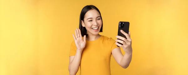 Immagine di felice, bella ragazza asiatica video chat, parlando su applicazione smartphone, in piedi su sfondo giallo — Foto Stock