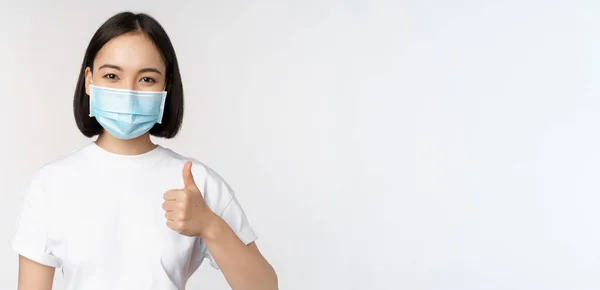 Le asiatisk flicka i medicinsk mask visar tummen upp, godkänna något bra, beröm och komplimang företag, står över vit bakgrund — Stockfoto