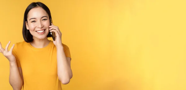 리언걸 이 휴대폰으로 얘기하고 있어. 스마트폰으로 전화하는 아시아 여성, 노란 배경 위에 서 있는 모습 — 스톡 사진