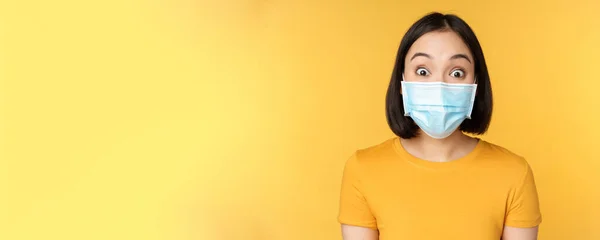 Ковид-19 и медицинская концепция. Крупным планом портрет азиатки в маске, удивлённой и удивлённой новостями, стоящей на жёлтом фоне — стоковое фото