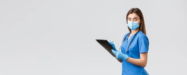 Covid-19, virüsün önlenmesi, sağlık, sağlık çalışanları ve karantina konsepti. Şok olmuş ve ürkmüş kadın hemşire ya da doktor mavi önlüklü, tıbbi maskeli, elinde pano bakışlarıyla kameraya şaşırmış.