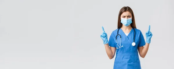 Covid-19, prevence viru, zdraví, zdravotnických pracovníků a koncepce karantény. Skeptická a zklamaná sestřička v modrých pláštích se cítí nespokojená, ukazuje prsty nahoru, nosí lékařskou masku — Stock fotografie