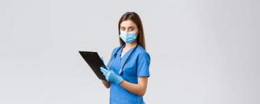 Covid-19, virüsün önlenmesi, sağlık, sağlık çalışanları ve karantina konsepti. Profesyonel kadın hemşire ya da doktor mavi önlüklü, tıbbi maske ve eldivenli hasta bilgilerini not alıyor.