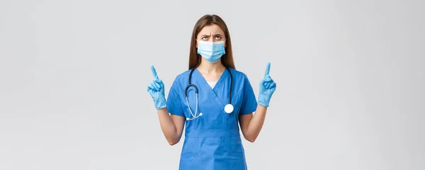 Covid-19, förebyggande av virus, hälsa, vårdpersonal och karantänkoncept. Orolig och besviken kvinnlig sjuksköterska eller läkare i blå skurar, medicinsk mask, rynka pannan tittar upp missnöjd — Stockfoto