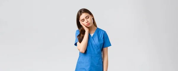 Healthcare workers, prevent virus and medicine concept. Tired sleepy female doctor, young nurse in blue scrubs, lean on palm and close eyes, exhausted from night shift at clinic fighting coronavirus — Stock Photo, Image