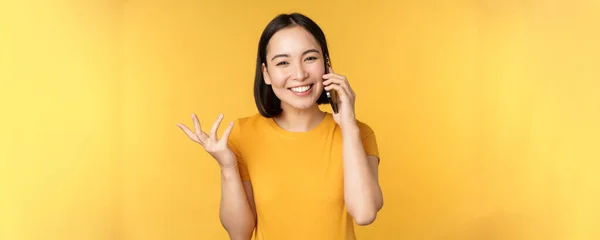 리언걸 이 휴대폰으로 얘기하고 있어. 스마트폰으로 전화하는 아시아 여성, 노란 배경 위에 서 있는 모습 — 스톡 사진