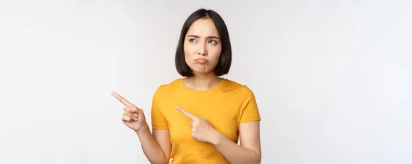 Ärgernis mürrisches asiatisches Mädchen, Schmollmund und Blick auf etliches unfair, Zeigefinger links auf Banner, Logo Marke, in gelbem T-Shirt vor weißem Hintergrund stehend — Stockfoto