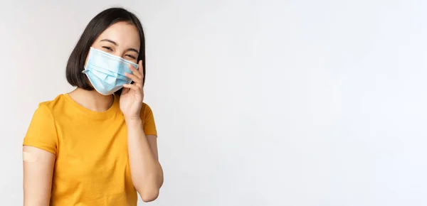 Covid-19, vaccinations- och sjukvårdskoncept. Söt asiatisk flicka i medicinsk ansiktsmask, visar band stöd efter coronavirus vaccination, stående över vit bakgrund — Stockfoto