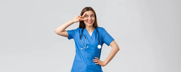 Pracownicy medyczni, opieka zdrowotna, koncepcja covid-19 i szczepień. Wesoły i energiczny młody lekarz, ładna pielęgniarka w niebieskim fartuchu, stetoskop, pokazać spokój w pobliżu oka i uśmiechnięty optymizm — Zdjęcie stockowe