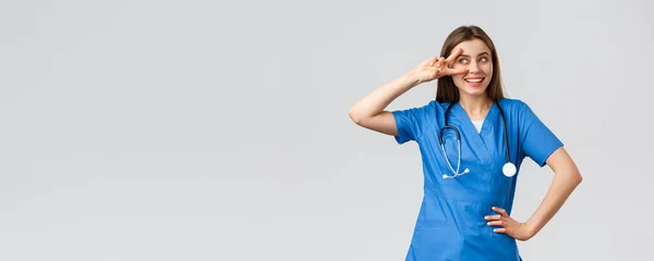 Travailleurs médicaux, santé, covide-19 et concept de vaccination. Joyeux et énergique jeune femme médecin, jolie infirmière dans les blouses bleues, stéthoscope, montrer la paix près des yeux et souriant optimiste — Photo