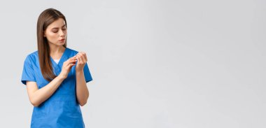 Sağlık çalışanları, virüsü, sigortayı ve ilaç konseptini engelleyin. Mavi önlüklü genç, güzel bir hemşire ya da doktor. Güzelliğini koruyor, tırnaklarına bakıyor, kuaför salonunu düşünüyor.
