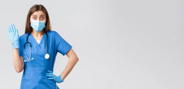 Covid-19, virüsün önlenmesi, sağlık, sağlık çalışanları ve karantina konsepti. Mavi önlüklü sevimli bayan hemşire merhaba diyor, klinikte birini görüyor, el sallıyor, maske takıyor ve eldiven takıyor.