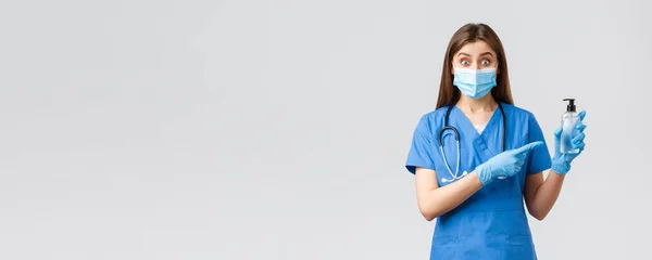Covid-19, förebyggande av virus, hälsa, vårdpersonal och karantänkoncept. Entusiastiska kvinnliga sjuksköterska eller läkare i blå skurar, medicinsk mask och handskar, pekar på hand desinfektionsmedel, ser förvånad — Stockfoto