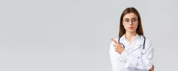 Healthcare workers, medicine, insurance and covid-19 pandemic concept. Serious-looking professional female doctor, nurse in white scrubs and glasses pointing finger left at banner or clinic info — Stock Photo, Image