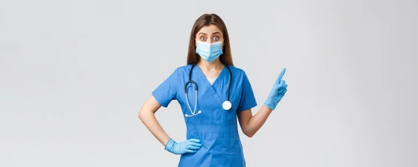 Covid-19, preventing virus, health, healthcare workers concept. Intrigued and excited female doctor, nurse in blue scrubs and medical mask inform patients, pointing fingers upper right corner — Stock Photo, Image