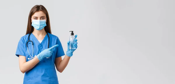 Ковид-19, профилактика вируса, здоровье, медицинские работники и карантин. Молодая профессиональная медсестра или врач в синих халатах, медицинская маска, указывающая пальцем на средство для дезинфекции рук — стоковое фото