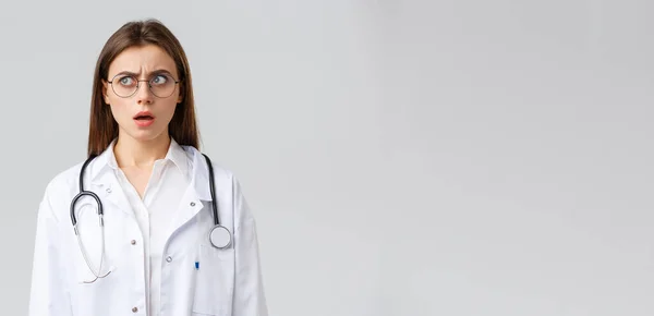 Healthcare workers, medicine, insurance and covid-19 pandemic concept. Shocked and confused young female doctor in white scrubs and glasses, stethoscope, stare left with concerned nervous face — Stock Photo, Image