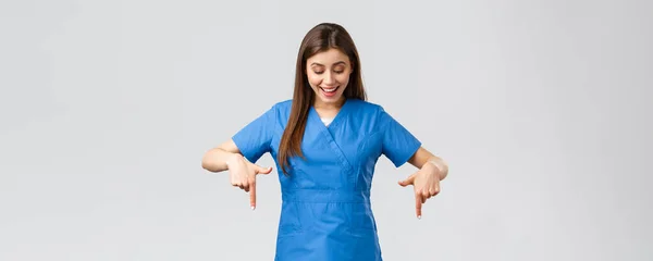 Healthcare workers, prevent virus, covid-19 test screening, medicine concept. Happy and excited pretty female nurse or doctor in blue scrubs, smiling as reading great news, pointing fingers down — Stock Photo, Image