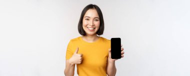 Teknoloji ve insan konsepti. Gülümseyen genç bir kadın başparmağını kaldırıyor ve akıllı telefon ekranı, cep telefonu uygulama arayüzü, beyaz arka plana karşı duruyor.