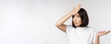 Kızgın Koreli kız facepalm, tokat alnı ve omuz silkme, pürüzsüzlükten kafası karışmış, beyaz arka planda duruyor.