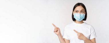 Tıbbî maske takmış Asyalı bir kadın, kafası karışmış sola bakıyor, logoyu işaret ediyor, reklamını yapıyor, beyaz arka planda duruyor.