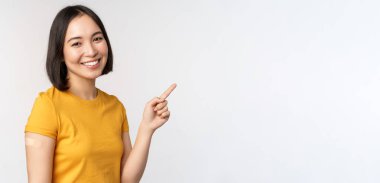 Covid-19 ve aşı konsepti. Shoudler 'a yara bandı yapıştırmış gülümseyen Asyalı kız, pankartı işaret ediyor, aşı kampanyasını gösteriyor, beyaz arka planda duruyor.