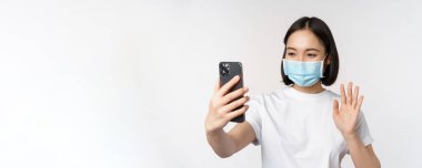 Sağlık ve covid-19 konsepti. Modern Asyalı kız, tıp maskeli öğrenci, cep telefonuyla görüntülü konuşma, akıllı telefon uygulamasına el sallama, beyaz arka planda dikilme