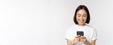 Gülümseyen Asyalı kadının portresi cep telefonu kullanıyor, sohbet ediyor, mesaj yazıyor, beyaz arka planda tişörtle dikiliyor.