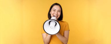 Gülümseyen Asyalı kadın megafonla duruyor, smth 'i anons ediyor, reklam ürünü, sarı arka planda duruyor.