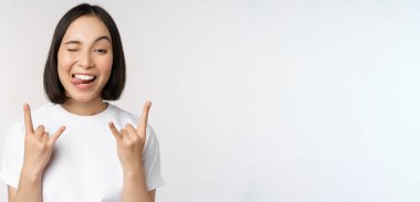 Şımarık Asyalı kız bağırıyor, konser veya festivalden zevk alıyor, rock 'ı gösteriyor, heavy metal işareti yapıyor, eğleniyor, beyaz arka planda duruyor.