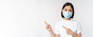 Medikal maskeli üzgün Asyalı kadın, parmaklarını sola gösteriyor, kaşlarını çatıyor ve üzgün görünüyor, şikayet ediyor, pankart gösteriyor, beyaz arka planda duruyor.