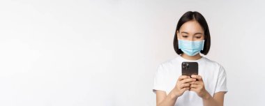Sağlık, ikna edici ve mobil konsept. Tıbbi maskeli genç Asyalı kadın, akıllı telefon ekranına bakıyor, telefon uygulaması kullanıyor, internetten alışveriş yapıyor, beyaz arka plan