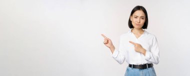 Şüpheci Asyalı kadının portresi somurtuyor, solu işaret ediyor ve şikayet ediyor, beyaz arka plana karşı durarak hoşnutsuzluğunu ya da hoşnutsuzluğunu ifade ediyor.