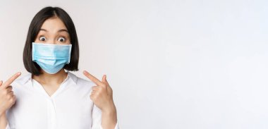 Tıbbî maske takıp beyaz arka planda duran Asyalı genç bir kadının görüntüsü.