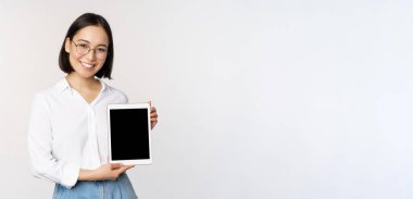 Genç Asyalı kadın profesyonel dijital tablet ekranı gösteriyor, cihazında bilgiler var, gülümsüyor ve kameraya bakıyor, beyaz arka planda duruyor.