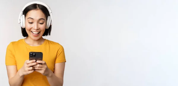 Söt japansk flicka i hörlurar, tittar på mobiltelefon och ler, med hjälp av musik app på smartphone, står mot vit bakgrund — Stockfoto